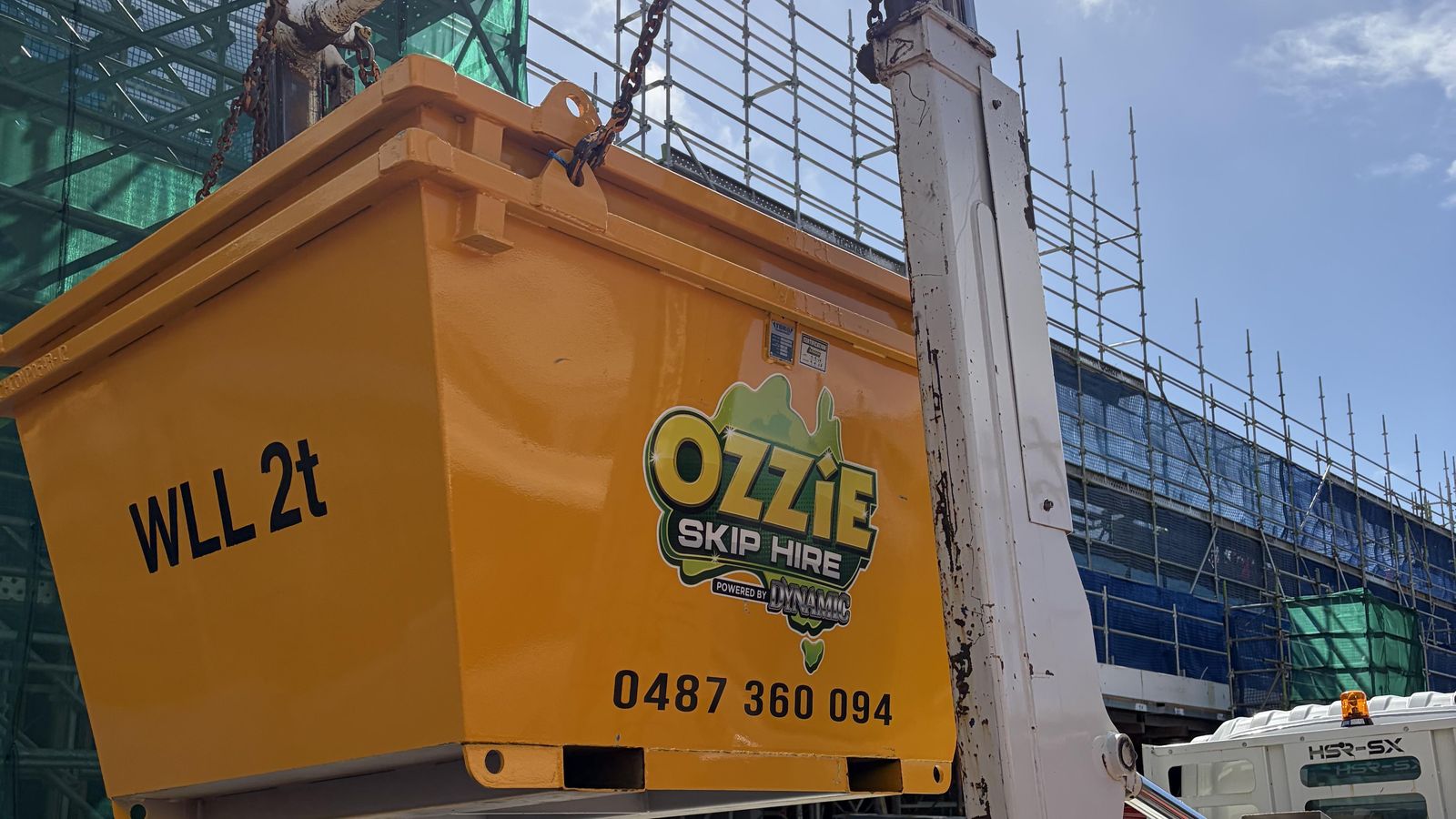 Ozzie Skip Hire crane-lift bin on the truck arms beside scaffolding at a Coffs Harbour construction site